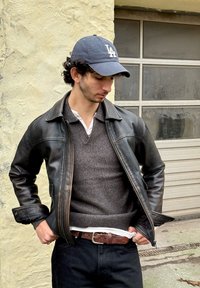 Veste en cuir noire avec un pull gris en dessous, jean foncé et casquette de baseball bleue. La veste est dotée d'une fermeture éclair et de poches latérales.