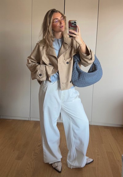 Trench beige con botones grandes, suéter gris claro, pantalones anchos blancos y un bolso azul texturizado. Zapatos de punta.