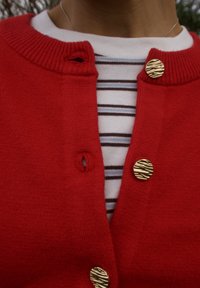 Cardigan rouge avec col côtelé, orné de deux boutons dorés à motifs. En dessous, un haut blanc rayé est partiellement visible.