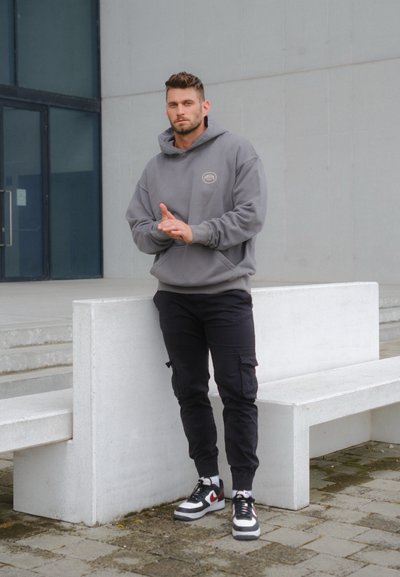 Sudadera gris con logo, pantalones cargo negros y zapatillas en blanco y negro. La persona está de pie frente a un fondo arquitectónico gris.