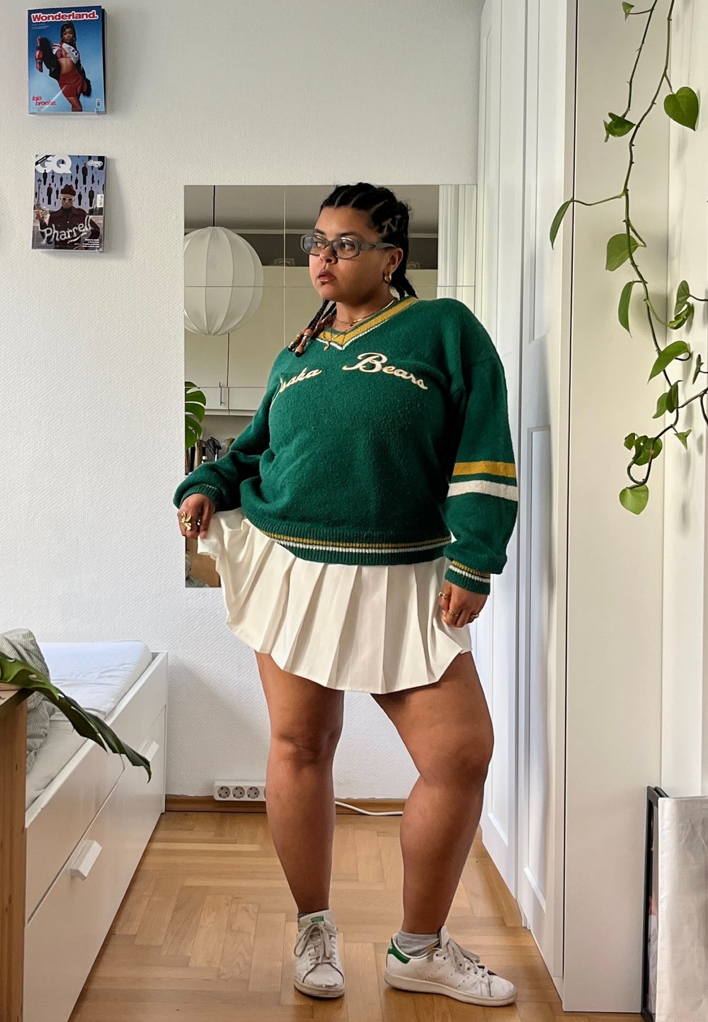 Πράσινο φλις πουλόβερ με κίτρινες και λευκές ρίγες, λευκή πτυχιστή φούστα και λευκά αθλητικά παπούτσια. Ο χώρος έχει ξύλινο πάτωμα και φυτά εσωτερικού χώρου.