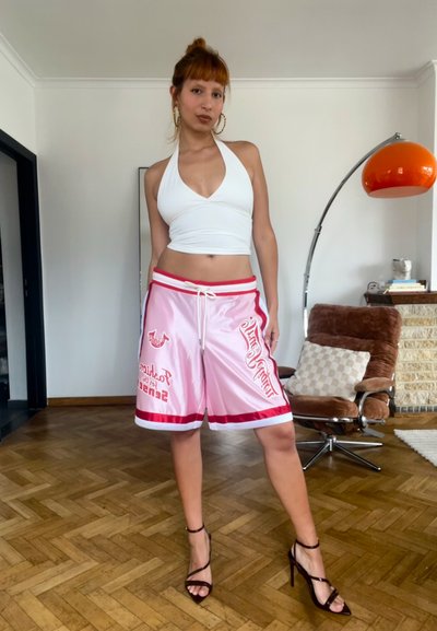 Mujer pelirroja con un top blanco de cuello halter, pantalones cortos de baloncesto rosas y tacones altos negros de pie sobre suelo de madera en un salón.