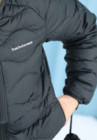 Veste matelassée noire avec le logo "Peak Performance" sur la poitrine, une main glissée dans la poche latérale, sur un fond bleu clair.