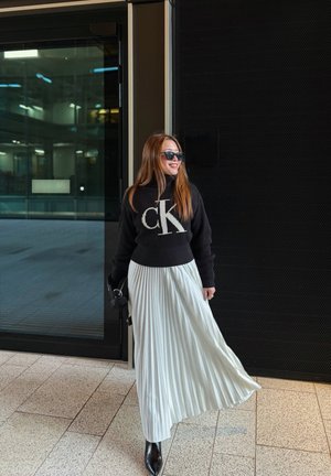 Jersey negro de punto con logo blanco, combinado con una falda plisada blanca hasta el suelo. Botas negras de punta afilada y gafas de sol. Textura lisa.