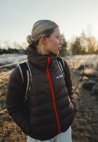 Jeune femme aux cheveux blonds attachés en chignon, portant une veste marron matelassée avec une fermeture éclair rouge, et un sac à dos, debout sur un chemin de terre près d'une clôture.