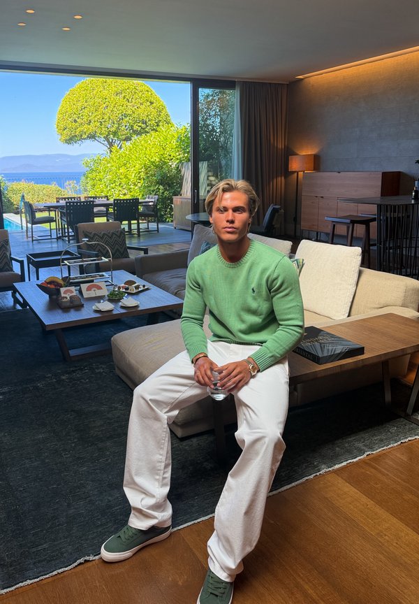 Jeune homme en pull vert et pantalon blanc assis sur un canapé dans un salon lumineux avec une porte vitrée donnant sur une terrasse extérieure et un jardin.
