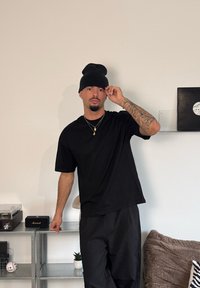 Svart t-shirt och svart mössa, står i en avslappnad pose. Enkel design, korta ärmar och enfärgad. Synliga tatueringar på ena armen.