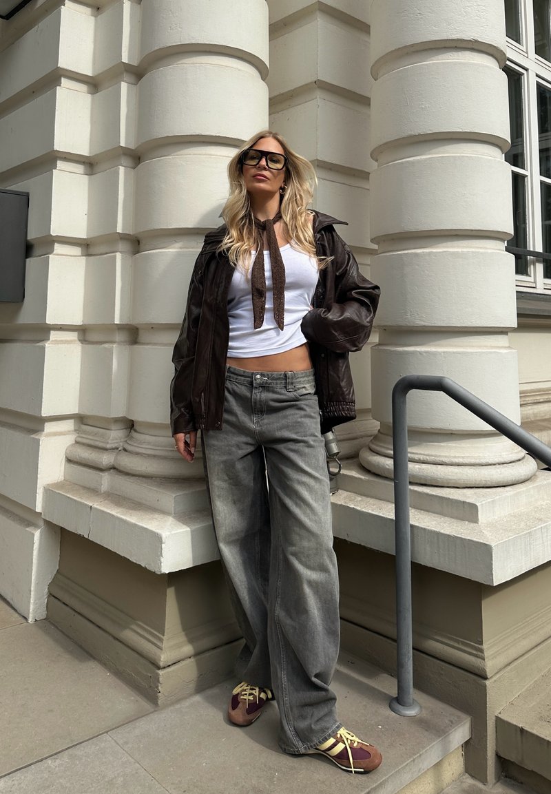 Giacca in pelle marrone, top bianco corto, jeans grigi a gamba larga e sneakers multicolori. Lunghi capelli biondi, in piedi contro un pilastro.