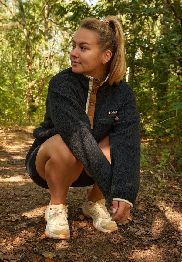 Fleece-pullover i mørk navy med krave og knapper i cream; lysebrune accenter; model, der squatter på en skovsti; iført lysfarvede sneakers.