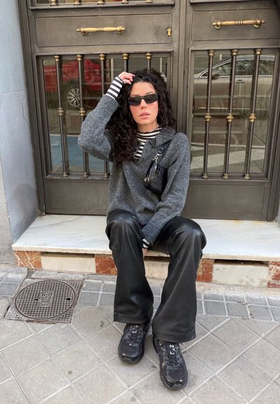 Mujer con cabello rizado y gafas de sol sentada en un escalón, con un suéter gris, camisa a rayas, pantalones negros, zapatillas y sosteniendo un bolso negro.