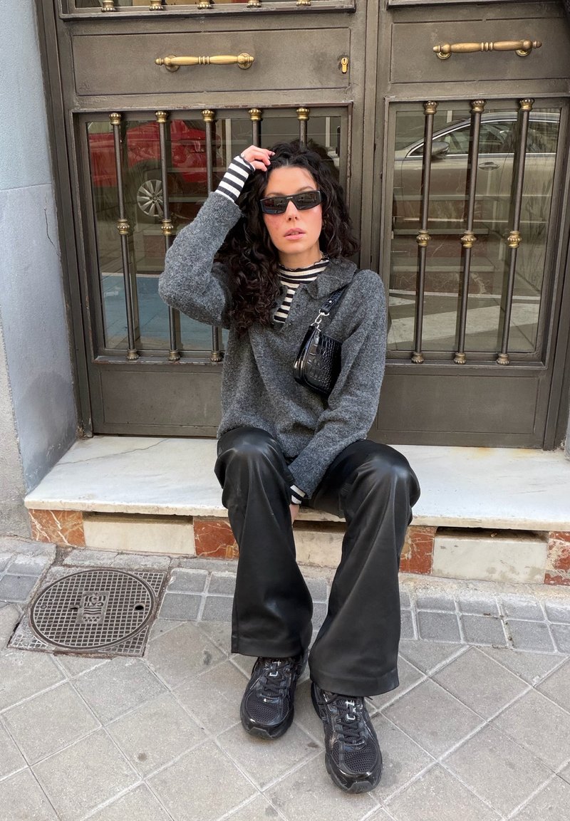 Donna con capelli ricci e occhiali da sole seduta su un gradino, indossa un maglione grigio, una camicia a righe, pantaloni neri, scarpe da ginnastica e tiene una borsa nera.