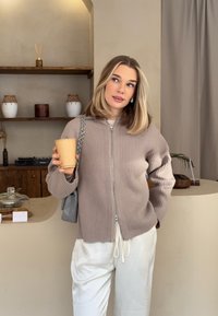 Femme blonde portant un pull à fermeture éclair beige et un pantalon blanc, tenant un gobelet en papier dans un café aux tons neutres avec des étagères et une décoration en bois derrière elle.