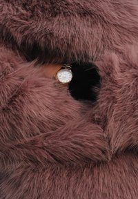 Blană artificială de culoare violet deschis înconjoară un ceas de mână auriu cu cadran alb și accente aurii, parțial vizibil prin textura blănii.