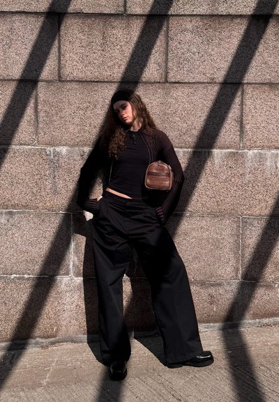 Pantalones negros de gran tamaño combinados con una blusa negra, un cárdigan a rayas marrón y un bolso marrón pequeño. El cabello rizado enmarca la cara.