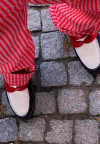 Scarpe in pelle bianca con dettagli blu navy e lacci rossi, abbinate a pantaloni a quadri rossi e bianchi, su pavimentazione in pietra testurizzata.
