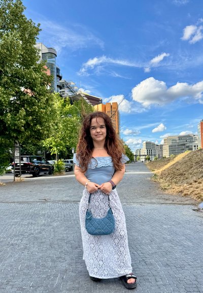 Top azul claro con detalles de encaje, falda maxi blanca de encaje y un bolso acolchado azul. Sandalias negras y cabello castaño ondulado en un entorno urbano al aire libre.