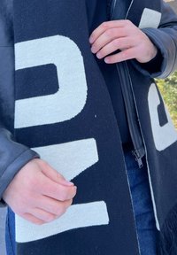 Zwart-witte gebreide sjaal met grote, gedurfde letters. Het materiaal lijkt zacht met een lichte textuur, franje aan de randen is zichtbaar.