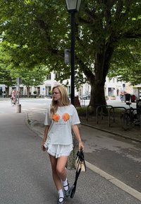 Camiseta branca com estampas de conchas laranja, combinada com calções de renda brancos. Mala preta e sapatos pretos com detalhes brancos. Ambiente urbano casual.