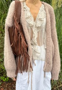 Cárdigan de punto color marrón claro con mangas oversized, superpuesto sobre una blusa blanca con volantes. Bolsa de cuero marrón con borlas. Falda blanca.