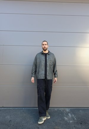 Chaqueta de mezclilla gris, camiseta negra, pantalones cargo negros, zapatillas verde oliva. De pie contra una pared gris claro, con un diseño minimalista.