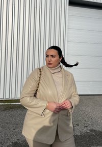 Μπεζ, γυαλιστερό δερμάτινο σακάκι πάνω από ένα ανοιχτό τζέρσεϊ με ψηλό λαιμό; συνδυασμένο με μία μικρή τσάντα χειρός με χρυσές αλυσίδες; φόντο από χαλίκι.