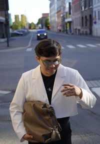 Ένα κομμένο λευκό σακάκι πάνω από ένα μαύρο πουκάμισο, κρατώντας μια καφέ δερμάτινη τσάντα. Οι ηλιό sunglasses είναι ανακλαστικοί, προσθέτοντας ένα μοναδικό γεωμετρικό σχήμα.