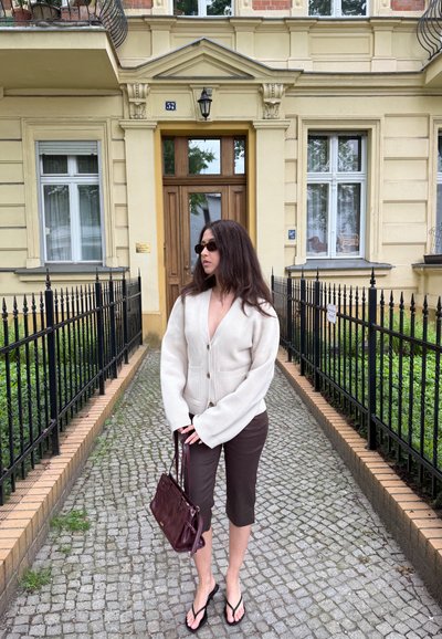 Mujer con gafas de sol, cárdigan blanco, pantalones cortos marrones y chanclas, de pie en un camino empedrado frente a la entrada de un edificio amarillo.
