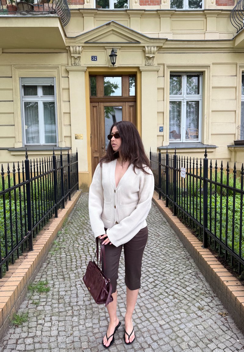 Mujer con gafas de sol, cárdigan blanco, pantalones cortos marrones y chanclas, de pie en un camino empedrado frente a la entrada de un edificio amarillo.