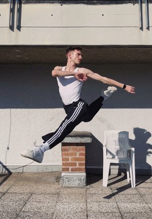 Hombre atlético vestindo una camiseta sin mangas blanca y pantalones de chándal negros con rayas blancas, ejecuta un salto sobre una superficie texturizada, con una silla al fondo.