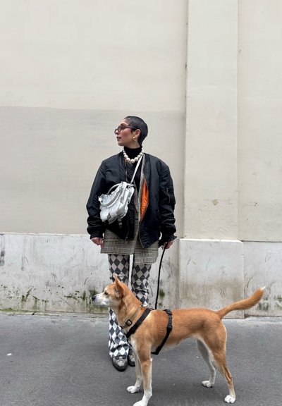 Persona vestida con una chaqueta bomber negra, una falda a cuadros y pantalones con patrón de diamantes, sosteniendo un bolso plateado. Un perro marrón claro con correa está a su lado.