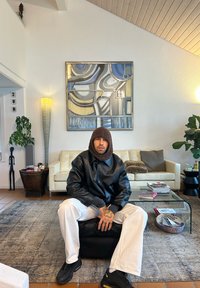 Osoba ubrana w czarną, skórzaną, oversize'ową kurtkę i brązowy dziergany kapelusz, siedząca na czarnej otomannie w nowoczesnym salonie z geometryczną sztuką na ścianie.