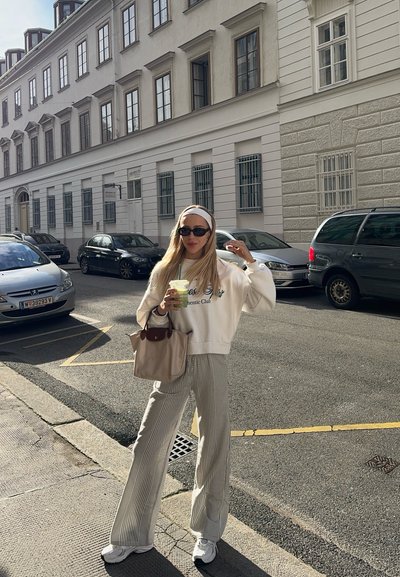 Sudadera blanca con texto verde, pantalones de pierna ancha a rayas y zapatillas blancas. Sosteniendo una bebida verde, de pie en una calle de la ciudad con coches.