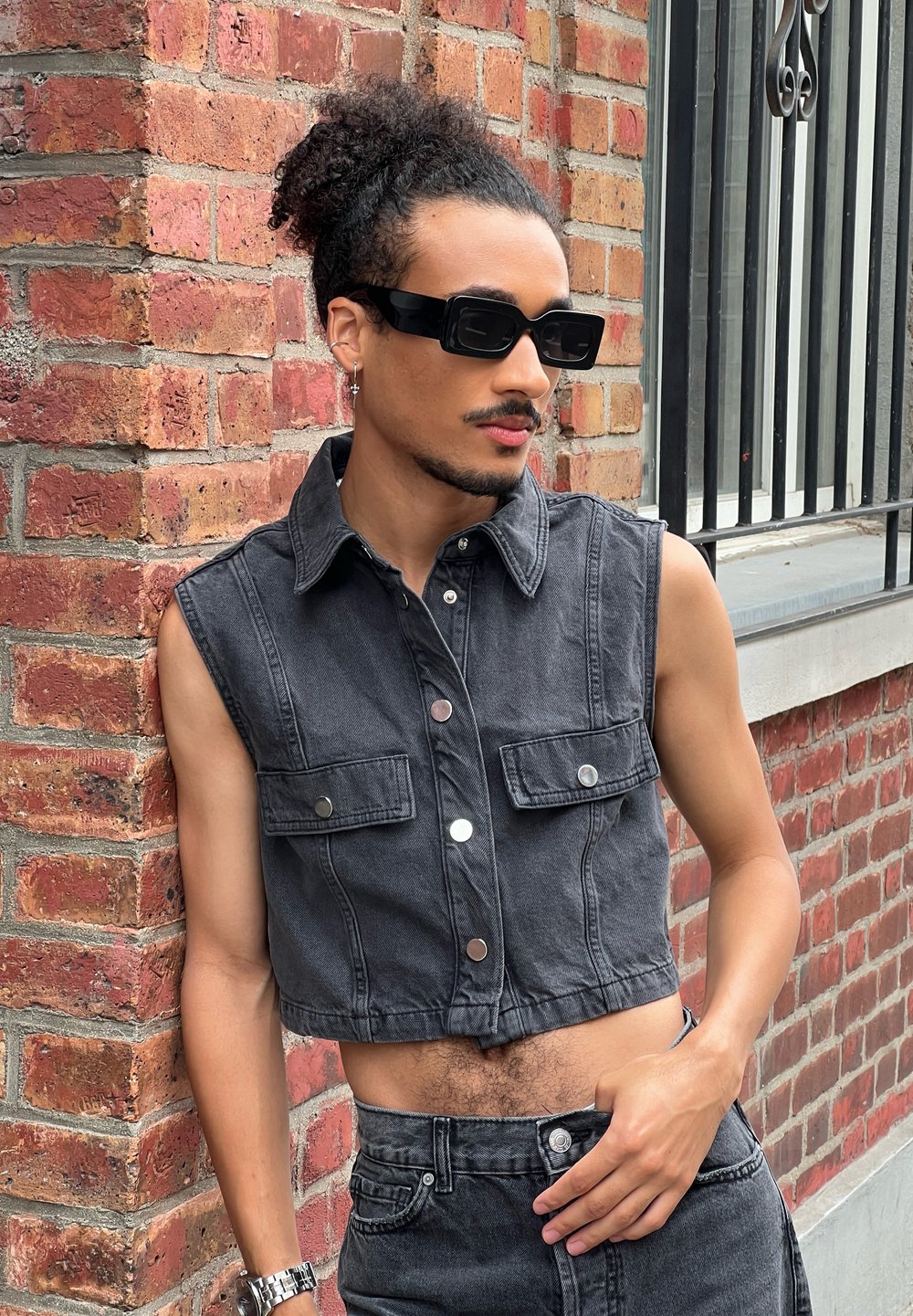 Skórzana czarna kamizelka z denimem, z dwoma przednimi kieszeniami, srebrnymi guzikami i kołnierzykiem, zestawiona z dopasowanymi spodniami o wysokim stanie na tle ceglanej ściany.