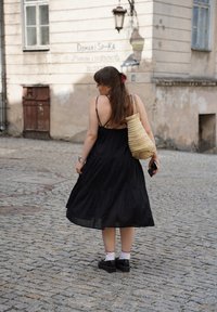 Abito nero a strati in tessuto leggero, indossato con calze bianche con bordo rosso e scarpe nere. Borsa in tessuto naturale a tracolla.