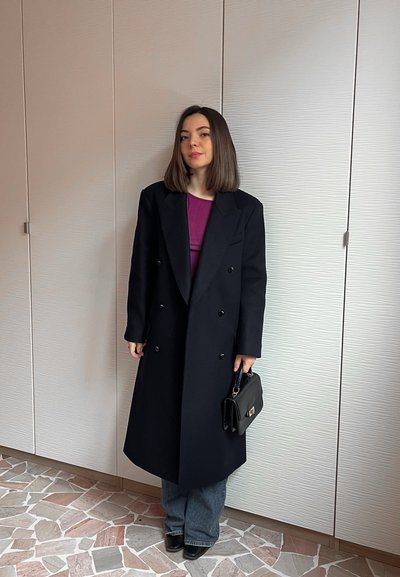 Joven con cabello castaño hasta los hombros, vestida con un abrigo largo negro, blusa morada, jeans azules, botas negras, sosteniendo un bolso negro en interiores.