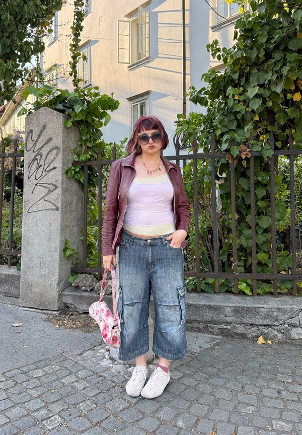 Person med kort rødt hår iført solbriller, vinrød skinnjakke, hvit topp, vide dongeribukser, hvite joggesko, holder en rosa blomstertaske ved et gjerde med grønt.