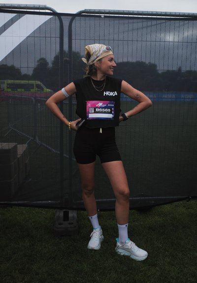 Camiseta sin mangas negra de HOKA, pantalones cortos negros, zapatillas blancas con acentos turquesa. Lleva una diadema con estampado y el número de carrera B6565 en el pecho.