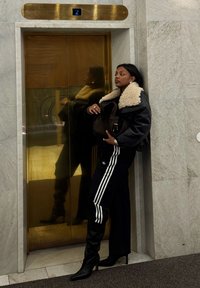 Kvinde i sort lammeskindsjakke og stribede bukser, der læner sig mod lukkede gyldne elevator-døre på anden sal.