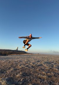 Figura atletica che salta indossando una felpa grigia e un berretto rosa, pantaloncini neri e sneakers multicolori su un prato ghiacciato, con il cielo blu.