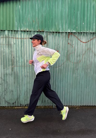 Mujer corriendo al aire libre con gorra negra, chaqueta blanca y amarilla, pantalones negros y zapatillas de correr de neón amarillo frente a una pared de metal corrugado verde.