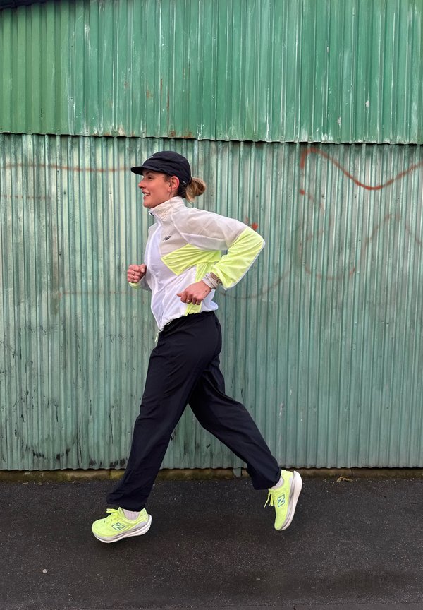 Vrouw die buiten aan het joggen is, gekleed in een zwarte pet, een witte en gele jas, zwarte broek en neongele hardloopschoenen, tegen een groene gegolfde metalen muur.