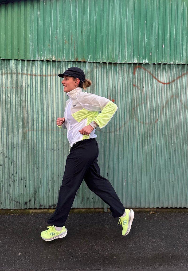 Vrouw die buiten jogt, gekleed in een zwarte pet, een wit en geel jasje, zwarte broek, en neongele hardloopschoenen, voor een groene gegolfde metalen muur.