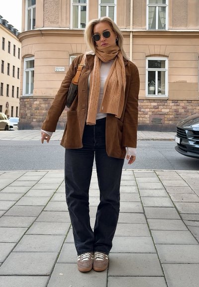 Blazer de ante marrón, bufanda de punto beige, camisa blanca, vaqueros negros, zapatillas marrones con cordones blancos, de pie sobre un pavimento de baldosas.