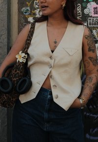 Beige ermeløs vest med fem knapper og en liten brystlomme, kombinert med mørke denimjeans og en leopardmønstret veske med hodetelefoner.