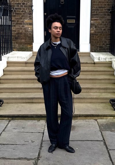 Persona con cabello rizado con una chaqueta de cuero negra, tops negros y blancos superpuestos, pantalones negros a rayas, zapatos negros, de pie cerca de escalones de piedra y una puerta.