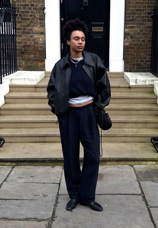 Person mit lockigem Haar in einer schwarzen Lederjacke, mehreren schwarzen und weißen Oberteilen, gestreiften schwarzen Hosen, schwarzen Schuhen, stehend in der Nähe von Steinstufen und einer Türöffnung.