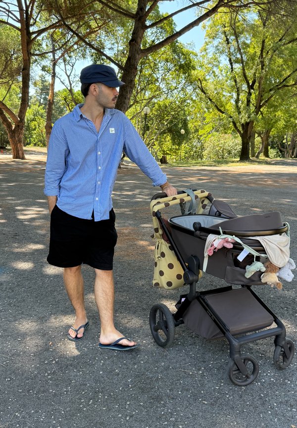 Homme en chemise bleue, short noir et casquette, debout sur un chemin à l'extérieur, tenant une poussette marron avec des jouets et un sac à pois.