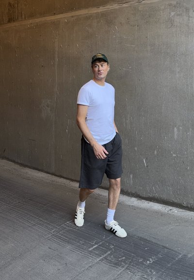 Hombre con camiseta blanca, pantalones cortos negros y zapatillas caminando frente a una pared de hormigón gris texturizada con una mano en el bolsillo.