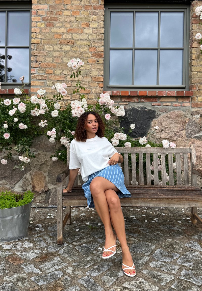 Femme aux cheveux bouclés portant un haut blanc et une jupe bleue assise sur un banc en bois avec des roses blanches et un mur de briques derrière elle.