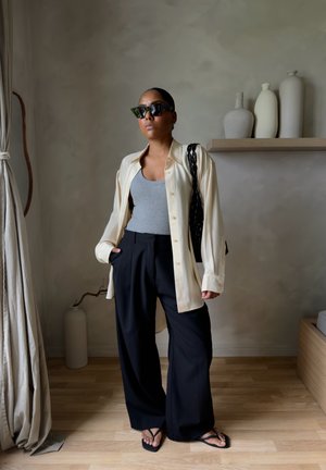 Mujer con gafas de sol negras, camisa crema oversize, camiseta gris, pantalones anchos negros y sandalias negras, de pie en el interior cerca de jarrones y cortinas.